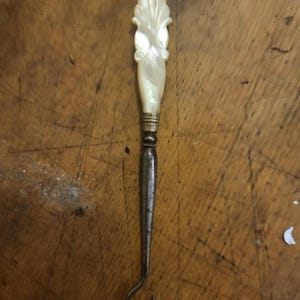 Pode incluir: Um gancho de botão antigo com uma alça decorativa de madrepérola. A alça apresenta um design floral esculpido. O gancho é feito de metal e tem uma extremidade curva. O gancho de botão é um item vintage.