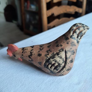 Könnte beinhalten: Eine Keramik-Hühnerfigur in einem gesprenkelten braun-schwarzen Muster, mit rotem Kamm und Kehllappen. Der Vogel liegt auf einer weißen Oberfläche und zeigt eine rustikale, handwerkliche Ästhetik. Die Figur ist ein dekoratives Objekt.