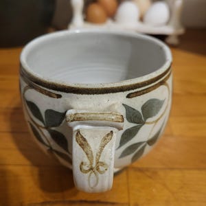May include: A ceramic soup bowl with a white interior and a cream-coloured exterior. The bowl is decorated with green leaves and brown accents. The handle features a decorative design. The bowl is sitting on a wooden surface.