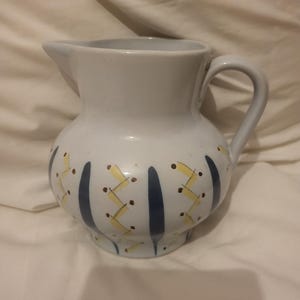 Op de afbeelding: Een witte keramische kan met een afgeronde body, tuit en handvat. De kan is versierd met verticale blauwe strepen en gele zigzagpatronen met bruine stippen. De kan wordt waarschijnlijk gebruikt voor het serveren van dranken.