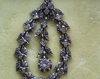 May include: An antique silver pendant with a teardrop design. The pendant is adorned with numerous small, clear gemstones and leaf-shaped accents. A large, round gemstone hangs at the bottom, adding a focal point to the piece. The pendant is displayed against a light yellow background.