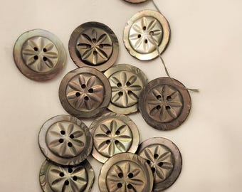 May include: A selection of iridescent, circular buttons featuring a floral design. Each button has four holes and a raised, star-shaped pattern in the centre. The buttons vary in colour, from silver to dark grey.