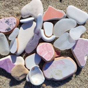 Puede incluir: Una colección de vidrio de mar liso y colorido y piedras de playa en tonos de rosa, blanco y marrón. El vidrio de mar es translúcido y tiene una variedad de formas y tamaños.