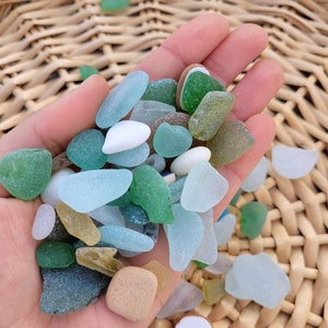 Puede incluir: Una mano sosteniendo una colección de vidrio de mar en varios tonos de azul, verde y blanco. El vidrio de mar es liso y pulido, con una variedad de formas y tamaños.