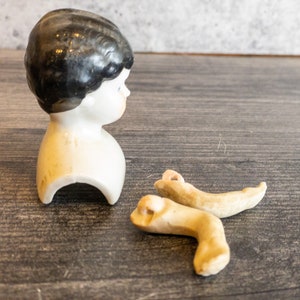 以下が含まれることがあります： 黒い髪の白い胸像のついた、磁器製のドールヘッド。磁器製のドールレッグも2本写っています。