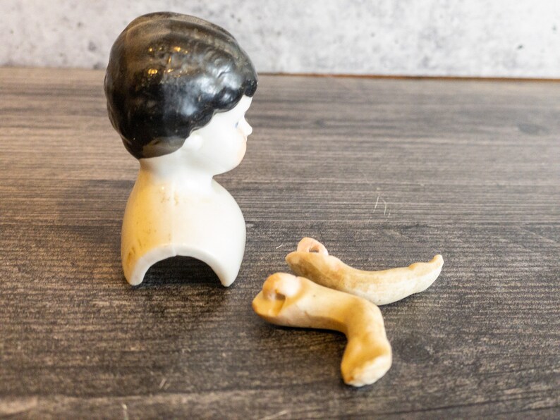以下が含まれることがあります： 黒い髪の白い胸像のついた、磁器製のドールヘッド。磁器製のドールレッグも2本写っています。
