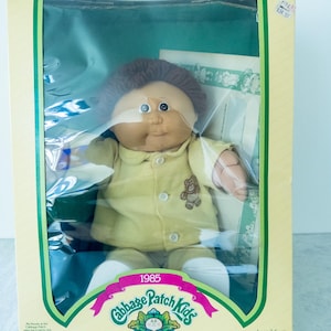 May include: A 1985 Cabbage Patch Kids doll with brown yarn hair and a tan complexion, wearing a yellow button-down shirt with a small bear graphic. The doll is enclosed in its original rectangular cardboard box with a clear plastic window front.