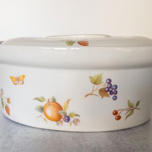 May include: White ceramic casserole dish with lid, decorated with colorful fruit and butterfly illustrations. The dish has a round shape with a lid and handles. The fruit illustrations include oranges, grapes, and berries.