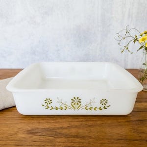 Puede incluir: Una fuente rectangular blanca para hornear con esquinas redondeadas y un diseño floral verde. La fuente está sobre una superficie de madera, con un paño beige y un pequeño jarrón con flores amarillas.