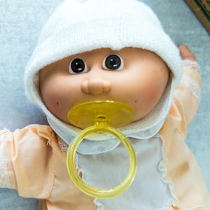 May include: A doll with a round face, dark brown eyes, and dimpled cheeks, wearing a white knit cap with a pom-pom. The doll has a yellow plastic pacifier in its mouth and is dressed in a light peach-coloured outfit with a white bib and lace trim.