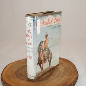 May include: A vintage hardcover book titled "The Hands of Cantu" by Tom Lea. The book cover features an illustration of a Native American man on horseback, riding through a desert landscape.