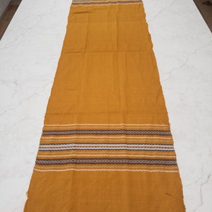 May include: A long, mustard yellow table runner with a woven pattern of white, blue and brown stripes. The runner has a fringe on both ends.