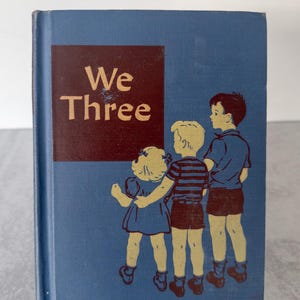 May include: Vintage children's book titled "We Three." The book has a blue cover with a maroon square containing the title in gold lettering. The cover features illustrations of three children in shorts and dresses.