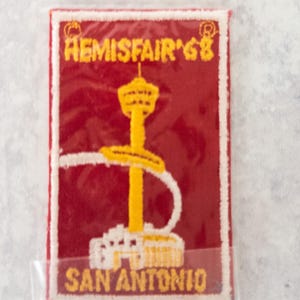 May include: A rectangular patch with a red background and white border. The patch features the text "HEMISFAIR '68" at the top and "SAN ANTONIO" at the bottom in yellow. A yellow tower and sword design is in the center.