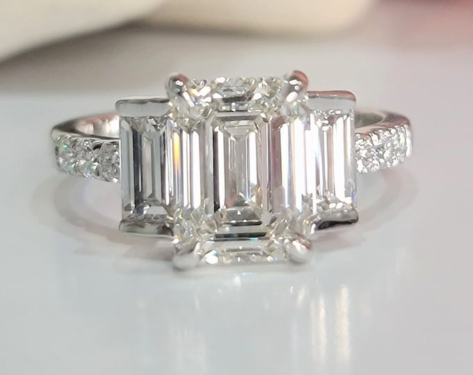 This Unique Timeless Three Stone Ring features a 3 Carat Emerald Cut IGI Certified CVD lab grown diamond accentuated with 0.70ct Step Cut Baguette side stones and 0.12 carats round brilliant diamonds