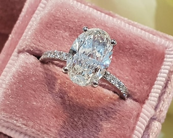 May include: A close-up of an oval-shaped diamond engagement ring set in a silver band. The ring is in a pink velvet ring box.