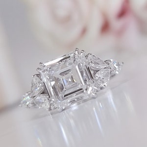 May include: A dazzling engagement ring featuring a large, square-cut center diamond, flanked by smaller, pear-shaped stones. The ring is set in a bright, polished metal, reflecting light. Soft, blurred pink flowers provide a delicate backdrop.
