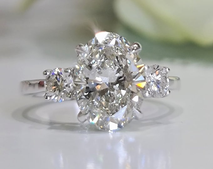 This Unique Three Stone Ring features a 3 Carat Oval Cut IGI Certified CVD lab grown diamond accentuated with 0.50ct round cut side stones
