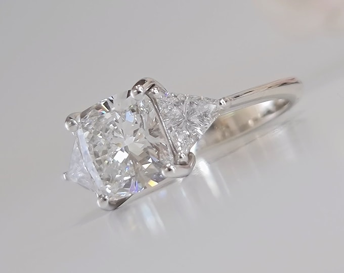 This Unique Three Stone Engagement Ring features a  2.5 Carat IGI Certified CVD lab grown Cushion Cut diamond accompanied by 0.70 carats triangle cut side diamonds