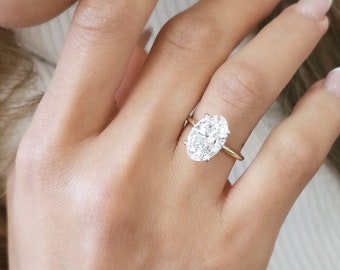 Może przedstawiać: Zbliżenie na dłoń z złotym pierścionkiem z dużym, owalnym diamentem. Diament jest otoczony mniejszymi diamentami.