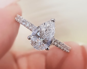 Puede incluir: Un anillo de compromiso de diamante de forma de pera con una configuración de pavé en la banda.