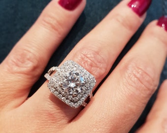 May include: A silver ring with a large square-shaped diamond surrounded by smaller diamonds. The ring is on a person's finger.