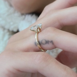 May include: A gold ring with a large, clear, pear-shaped diamond. The ring is worn on the finger of a person with a tattoo that says "MOM".