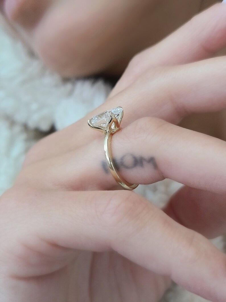 May include: A gold ring with a large, clear, pear-shaped diamond. The ring is worn on the finger of a person with a tattoo that says "MOM".