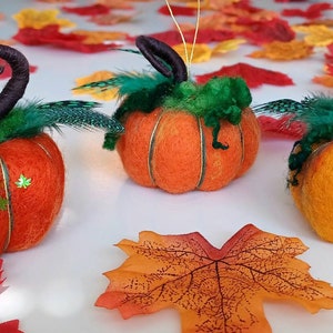 May include: Three orange felt pumpkins with green felt leaves and stems. The stems are curled and have black feathers attached. The pumpkins are sitting on a white surface with autumn leaves scattered around them.