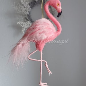 Könnte beinhalten: Ein rosafarbener Filz-Flamingo-Anhänger mit langem Hals und dünnen Beinen. Der Flamingo steht auf einem Bein und hat ein schwarzes Auge und einen schwarzen Schnabel.