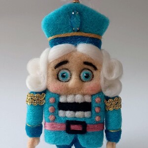 Könnte beinhalten: Ein Nussknacker-Anhänger aus Filz mit blauer Uniform, weißen Haaren und einer rosa Feder auf seinem Hut. Er hat ein breites Lächeln und große blaue Augen.