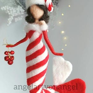 Könnte beinhalten: Ein Filz-Weihnachtsornament in Form eines Zuckerstangs mit einem weiß-rot gestreiften Körper, weißen Flügeln und einem weißen Hut mit einem roten Zuckerstang oben drauf. Das Ornament hält einen Bund roter Glöckchen.
