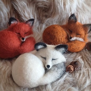 Könnte beinhalten: Drei Filzfiguren von Füchsen, eine orange, eine braun und eine weiß mit grauen Akzenten, die auf einer Kunstfell-Oberfläche zusammengerollt schlafen.