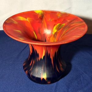 May include: A red and black glass vase with a wide, flared opening and a swirling, abstract design. The vase has a dark blue base and a bright red top with yellow and orange accents.