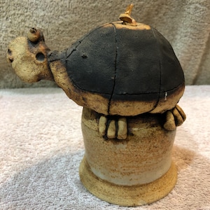 May include: A ceramic turtle figurine with a black shell and a beige body. The turtle is perched on a beige ceramic base.