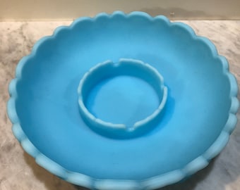 Könnte beinhalten: Eine hellblaue Schale aus satiniertem Glas mit gewelltem Rand und einer mittig erhöhten, kreisförmigen Vertiefung. Das Objekt besitzt eine glatte, matte Oberfläche und eine durchgehend einheitliche, opake blaue Farbe, die auch den gewellten Rand umfasst.