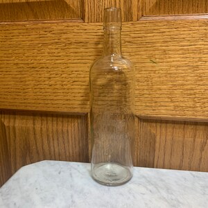 May include: A clear glass bottle with a long neck and a rounded base. The bottle is empty and appears to be vintage. The background is a wooden door and a white marble surface.
