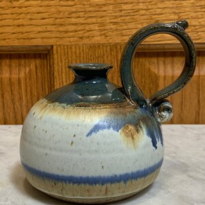 May include: Handmade ceramic pitcher with a rounded body and a large handle. The pitcher features a color palette of white, blue, and brown. The handle is arched and attached to the top and side of the pitcher. The pitcher has a small spout.
