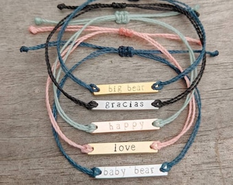 Op de afbeelding: Vijf kleurrijke vriendschapsarmbanden met metalen staafbedels. De bedels hebben de woorden "big bear", "gracias", "happy", "love" en "baby bear" erop gegraveerd.