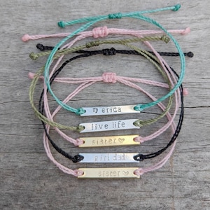 May include: A set of six colorful friendship bracelets with personalized metal tags. The tags have the following text: "my erica", "live life", "sister", "girl dad", and "siscer".