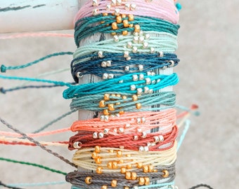 Könnte beinhalten: Ein Stapel bunter Armbänder mit goldenen und silbernen Perlen. Die Armbänder sind aus Schnur und zu einem Bündel zusammengebunden.