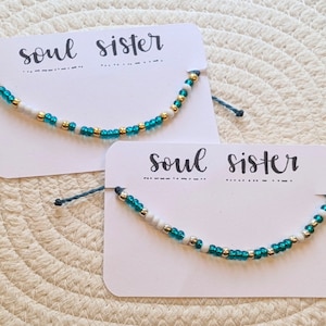 Puede incluir: Dos tarjetas blancas con el texto "soul sister" escrito en cursiva negra. Cada tarjeta tiene una pulsera de cuentas azul verdoso y blanca atada.