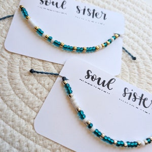 May include: Two white cards with the text "soul sister" written in black ink. Each card has a teal and white beaded bracelet with gold accents.