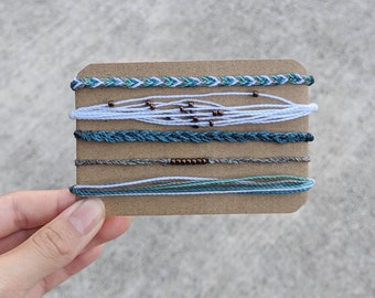 May include: A set of six colorful friendship bracelets with a mix of white, blue, and gray threads. The bracelets are displayed on a brown card.