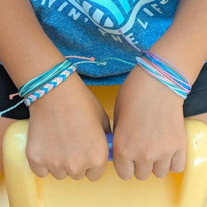 Puede incluir: Dos pulseras de amistad hechas a mano en tonos de azul, rosa y blanco son visibles en las muñecas. Una pulsera presenta un diseño trenzado, mientras que la otra está hecha de múltiples hebras. Las pulseras están en las muñecas de una persona.