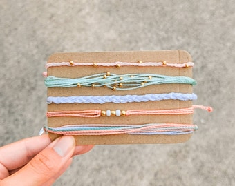 Op de afbeelding: Een set van zes kleurrijke vriendschapsarmbanden met gouden kralen en witte kralen. De armbanden worden gepresenteerd op een bruine kaart.