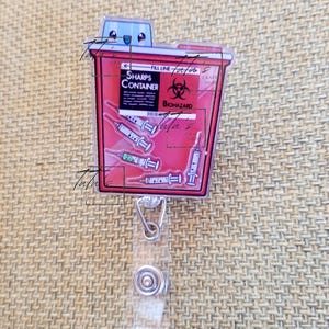 May include: A red and white badge reel featuring a cartoon sharps container design. The container has the words "SHARPS CONTAINER" and "BIOHAZARD" printed on it, along with syringe illustrations. The reel has a clear plastic strap and a metal clip.