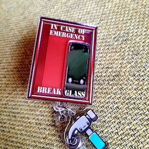 May include: A novelty badge reel with a red and black frame that reads "IN CASE OF EMERGENCY BREAK GLASS." The reel features a cartoon smartphone graphic and a hammer charm with a blue handle. The reel is attached to a clear plastic clip.