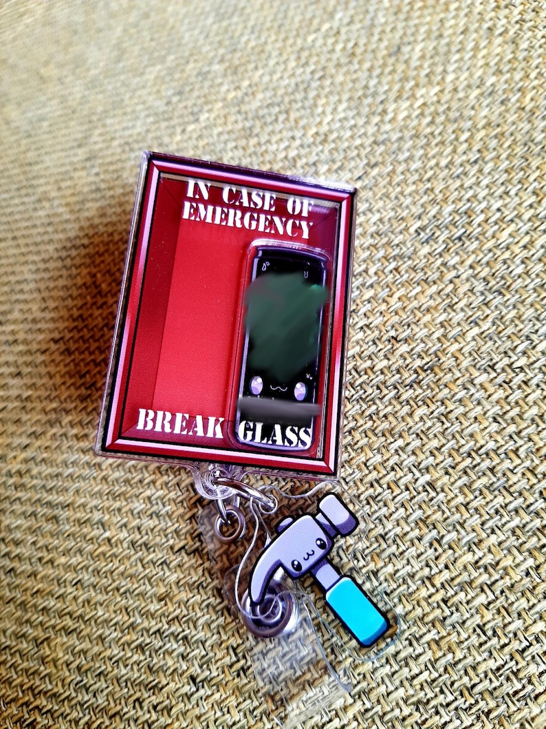 May include: A novelty badge reel with a red and black frame that reads "IN CASE OF EMERGENCY BREAK GLASS." The reel features a cartoon smartphone graphic and a hammer charm with a blue handle. The reel is attached to a clear plastic clip.