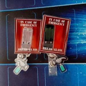 May include: Two novelty badge reels with the text "IN CASE OF EMERGENCY" and "BREAK GLASS". One reel features a clear glass design, the other a green glass design. Each has a hammer charm with a teal handle.